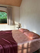 A cozy bedroom featuring Rimmba’s Cosmic Galaxy Pink Full Silk Bedding Set with two matching pillows, a burgundy throw, a side table and ceramic lamp, beige curtains at the window, and organic plants outside.