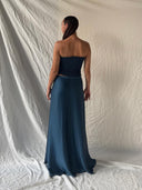 A woman faces away from the camera, wearing the Indigo Silk Dayu - Elastic Bandeau Top by Rimmba | Sustainable Clothing and a matching skirt, standing before a light, wrinkled fabric backdrop.