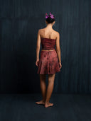 Barefoot woman wearing a silk red ecoprint strapless top and mini silk wrap skirt against a dark background.
