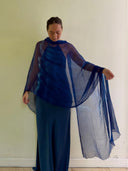 A person stands against a plain light wall, wearing the Deep Blue - Indigo silk scarf by Rimmba | Sustainable Clothing—handcrafted from mulberry silk and tinted with natural plant dyes—looking down and holding out one side gracefully.