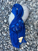 The Deep Blue - Indigo silk scarf by Rimmba | Sustainable Clothing, crafted from Mulberry Silk and plant-dyed, is draped over an oval gray stone with pebbles around it. A beige tag on the scarf highlights product details.