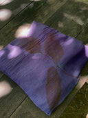 The Eco-Print Silk Chiffon Scarf - Purple by Rimmba | Sustainable Clothing, featuring a leaf print from botanical contact printing, rests on weathered wood as dappled sunlight casts light and shadow across its Mulberry silk surface.