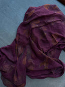 The Eco-Print Silk Chiffon Scarf - Magenta Purple by Rimmba | Sustainable Clothing features sheer, purple silk chiffon with subtle brown botanical prints, elegantly draped on a blue textured fabric surface.