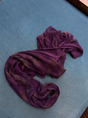 The Rimmba Eco-Print Silk Chiffon Scarf in Magenta Purple is loosely arranged in an abstract shape on a light blue fabric surface, illuminated by natural light from the top right.