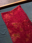 The Eco-Print Silk Chiffon Scarf - Red by Rimmba | Sustainable Clothing features subtle orange botanical patterns on deep red silk chiffon, displayed on a dark gray surface beside a wooden edge, highlighting its light, slightly wrinkled texture.