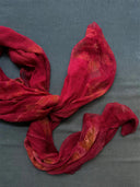 The Eco-Print Silk Chiffon Scarf - Red by Rimmba | Sustainable Clothing is a sheer, crinkled silk scarf with golden patterns, artfully arranged on a textured grey surface, showcasing the beauty of botanical contact printing.