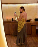 Karunia Fischer's taking mirror selfie in RIMMBA  store. She is wearing ecoprint marigold scarf as a top and paired with a long, gray skirt. Her long, dark hair is worn down, and she is turned slightly sideways, looking at her phone screen while capturing the photo.