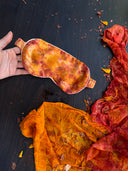 A hand holds Rimmba's Eco-Print Silk Eye-Mask, an anti-aging sleep accessory with vibrant orange and red tie-dye. Below, botanically dyed fabric adorned with petals creates a dark, autumnal surface effect.
