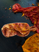 The Eco-Print Silk Eye-Mask by Rimmba | Sustainable Clothing, featuring a marbled orange and red pattern from botanical dyes, rests on a dark surface amid matching fabric scraps and shavings, creating a vibrant and textured scene.
