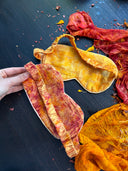 A hand displays two Eco-Print Silk Eye-Masks by Rimmba | Sustainable Clothing on a dark surface, surrounded by vibrant, crumbled fabric in shades of red, orange, and yellow with scattered dried flower petals.
