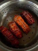 Four cylindrical packages, resembling food items, are in a metal steamer basket. Tied with string and boasting a reddish-brown hue, they appear botanically dyed, akin to the Eco-Print Silk Eye-Mask by Rimmba | Sustainable Clothing.