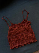 A rust-colored Frida Crinkle Crop-Top by Rimmba | Sustainable Clothing, made from 100% Mulberry Silk, with ruching, thin spaghetti straps, and a ruffled hem is laid flat on a dark textured surface.
