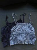 Two Frida Crinkle Crop-Tops by Rimmba | Sustainable Clothing—one dark gray, one light silver-gray—crafted from 100% Mulberry Silk, with thin straps, are laid flat on dark fabric. Part of our Zero Waste program.