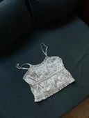 The Frida Crinkle Crop-Top by Rimmba | Sustainable Clothing, a white sleeveless top with thin straps and ruffled details, is displayed on a dark blue cushioned surface.