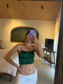 A person takes a mirror selfie in a cozy, sunlit bedroom, wearing a green Frida Crinkle Crop-Top by Rimmba | Sustainable Clothing and a white wrap skirt. The room features neutral decor, pottery accents, and a large window.