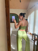 A woman stands in a sunlit room, facing away from the mirror while taking a photo with her phone. She wears a light green Frida Crinkle Crop-Top by Rimmba | Sustainable Clothing and matching pants, her hair tied in a bun.