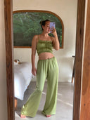 A woman takes a mirror selfie wearing the Frida Crinkle Crop-Top by Rimmba | Sustainable Clothing, paired with matching wide-leg pants. A bed and a window with greenery are visible in the background.