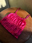 A Frida Crinkle Crop-Top by Rimmba | Sustainable Clothing, in hot pink ruched satin with spaghetti straps, made from 100% Mulberry Silk, is displayed on a round wooden table, sunlit.