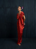A woman stands barefoot against a dark textured background, wearing the loose-fitting Gaya Silk Kaftan Dress - Ecoprint Red by Rimmba | Sustainable Clothing. She has a purple flower in her hair, eyes closed, and hands behind her back.