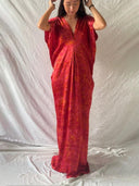 A woman stands before a white sheet backdrop wearing the Gaya Silk Kaftan Dress - Ecoprint Red by Rimmba | Sustainable Clothing, a loose, flowing v-neck kaftan with short sleeves, her hands in her hair as she looks downward.