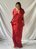 A person with straight, dark hair stands barefoot on white fabric, wearing the Gaya Silk Kaftan Dress - Ecoprint Red by Rimmba | Sustainable Clothing, gently holding the sides and looking down against a white sheet backdrop.