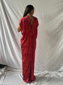 A person with long dark hair, seen from behind, wears the Gaya Silk Kaftan Dress - Ecoprint Red by Rimmba | Sustainable Clothing. The loose, floor-length dress has short sleeves and is set against a plain, wrinkled white backdrop.