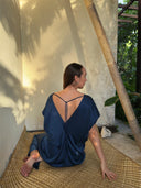 A woman in the Gaya Silk Kaftan Dress - Indigo by Rimmba | Sustainable Clothing sits on a woven mat, her back to the camera. Sunlight and leafy shadows create a peaceful scene inspired by botanical dyes.
