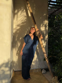 A woman in the Gaya Silk Kaftan Dress - Indigo from Rimmba | Sustainable Clothing stands barefoot on a mat, leaning against a bamboo pole as sunlight and leafy shadows play around her, enjoying the natural outdoor setting and botanical dyes.