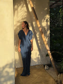 A woman in the Gaya Silk Kaftan Dress - Indigo by Rimmba | Sustainable Clothing stands barefoot on a woven mat, leaning against a sunlit wall with leafy shadows, surrounded by greenery and inspired by botanical dyes.