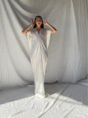 A woman in the Gaya Silk Kaftan Dress - White by Rimmba | Sustainable Clothing stands before draped white fabric, eyes closed and hands near her head, embodying a serene and ethereal mood.