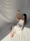 A woman in the Gaya Silk Kaftan Dress - White by Rimmba | Sustainable Clothing kneels on a fabric-draped floor, facing sideways with one arm raised as soft side light highlights her. The background features flowing white fabric.