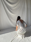 A person with long brown hair kneels on a fabric-covered floor, facing away, wearing the Rimmba Gaya Silk Kaftan Dress - White, as soft light and shadows play across flowing white fabric in the background.