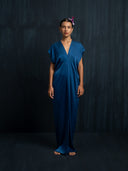 A woman stands barefoot against a dark textured background, wearing the Rimmba Gaya Silk Kaftan Dress in indigo, featuring short sleeves and a V-neck. Her slicked-back hair is adorned with a purple flower.