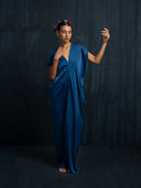 A woman stands barefoot against a dark backdrop, eyes closed and one arm raised, wearing the Gaya Silk Kaftan Dress in indigo by Rimmba | Sustainable Clothing, adorned with purple flowers in her hair.