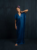 A woman stands barefoot against a dark background in the Gaya Silk Kaftan Dress - Indigo by Rimmba | Sustainable Clothing, tilting her head with a pink flower in her sleek hair and resting one hand on her neck.