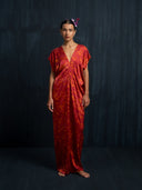 A woman stands barefoot against a dark background, wearing the Gaya Silk Kaftan Dress – Ecoprint Red by Rimmba | Sustainable Clothing. Her hair is styled back with a purple flower accessory.