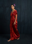 A woman stands barefoot against a dark textured background, wearing the Gaya Silk Kaftan Dress - Ecoprint Red by Rimmba | Sustainable Clothing, adorned with purple flowers in her hair and a serious expression.