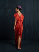 A woman stands barefoot against a dark backdrop, wearing the Rimmba Gaya Silk Kaftan Dress in Ecoprint Red. Her hair is styled in an updo with purple flowers as she looks over her shoulder.