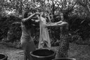 The black-and-white image shows two women standing outdoors, each holding a piece of fabric that appears to be wet.The setting is natural, surrounded by trees and plants, suggesting that they are engaged in a traditional or artisanal fabric-dyeing activity in a garden or outdoor workspace.