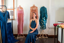 A woman in a blue dress sits on a chair, smiling, surrounded by colorful dresses displayed on mannequins and racks in a bright, modern boutique.
