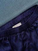 Close-up of the waistband of His & Hers Silk Bottoms - Boxers in dark blue by Rimmba | Sustainable Clothing, featuring a white “RIMMA” label and crafted with a zero waste design, displayed on a textured teal surface.