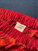 Close-up of the waistband on Rimmba’s His & Hers Silk Boxers Bottoms – Klimt, in a red-orange pattern with a "RIMMBA 100% SILK" fabric label, set on a dark blue textured background. Rimmba | Sustainable Clothing.