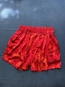 A pair of "His & Hers Silk Boxers Bottoms - Klimt" by Rimmba | Sustainable Clothing, featuring a textured, slightly crinkled red mulberry silk and elastic waistband, is displayed on a dark blue fabric background.