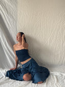 A woman wears the Indigo Silk Dayu - Elastic Bandeau Top by Rimmba | Sustainable Clothing, sitting on the floor with eyes closed and one hand in her hair against draped white fabric.
