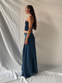 A woman with long brown hair stands in profile against a white textured backdrop, wearing a dark blue strapless top and the Jannah Indigo - Wrap Skirt by Rimmba | Sustainable Clothing, crafted from mulberry silk and botanical dyes.