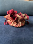 The Rimmba | Sustainable Clothing Jewelry Pouch, crafted from shiny red and gold satin with a secure drawstring, rests on a blue upholstered surface, its top ruffled open.