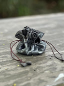 The Rimmba | Sustainable Clothing Jewelry Pouch, made of shiny gray satin with ruffled edges and secure brown drawstrings, rests on a weathered wood surface with a softly blurred background.