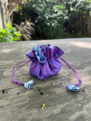 The Rimmba | Sustainable Clothing Jewelry Pouch, featuring purple satin with blue accents and a secure drawstring, rests on a weathered outdoor wooden table surrounded by lush green plants.