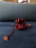 The Rimmba | Sustainable Clothing Jewelry Pouch, crafted in shiny red satin with a secure drawstring closure, rests on a dark blue surface against a softly blurred blue and beige background.