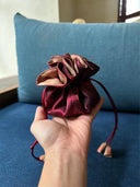 A hand holds the Rimmba | Sustainable Clothing Jewelry Pouch—small, shiny maroon satin with gold accents and a secure drawstring closure—set against a blue cushion background.